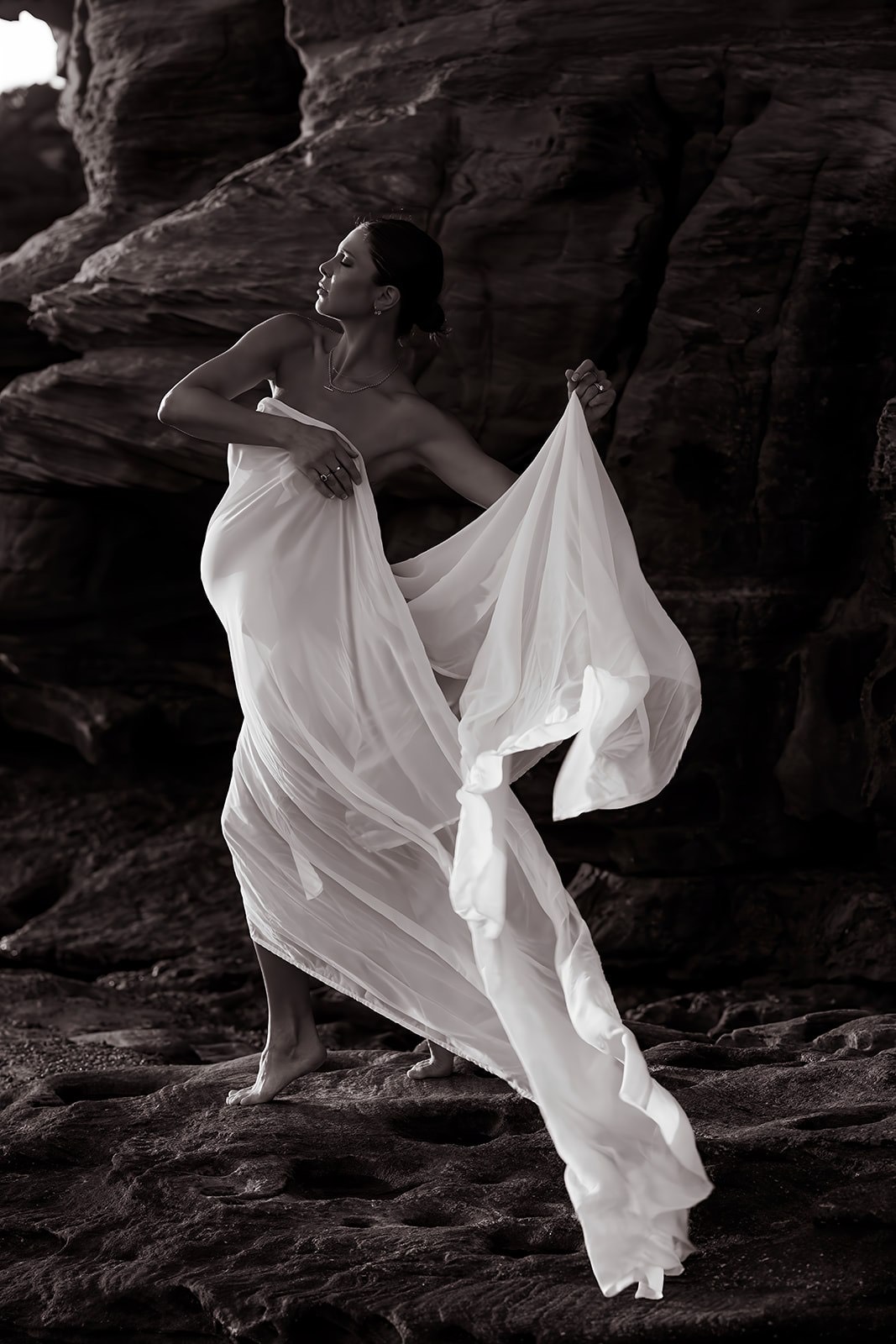 Sculptural maternity portrait moving with white fabric among coastal rocks