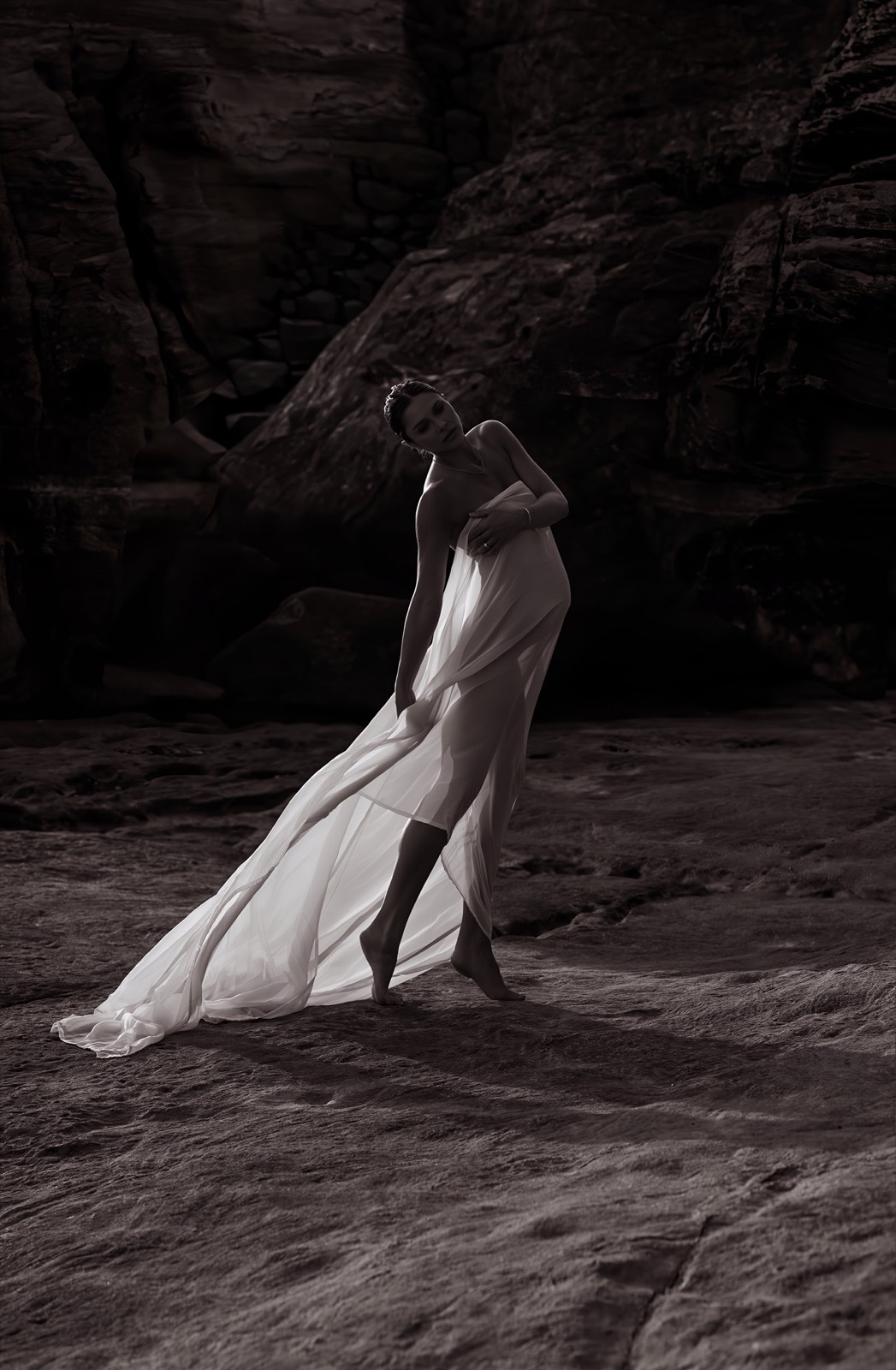 Distant maternity portrait in flowing white fabric against dark sandstone